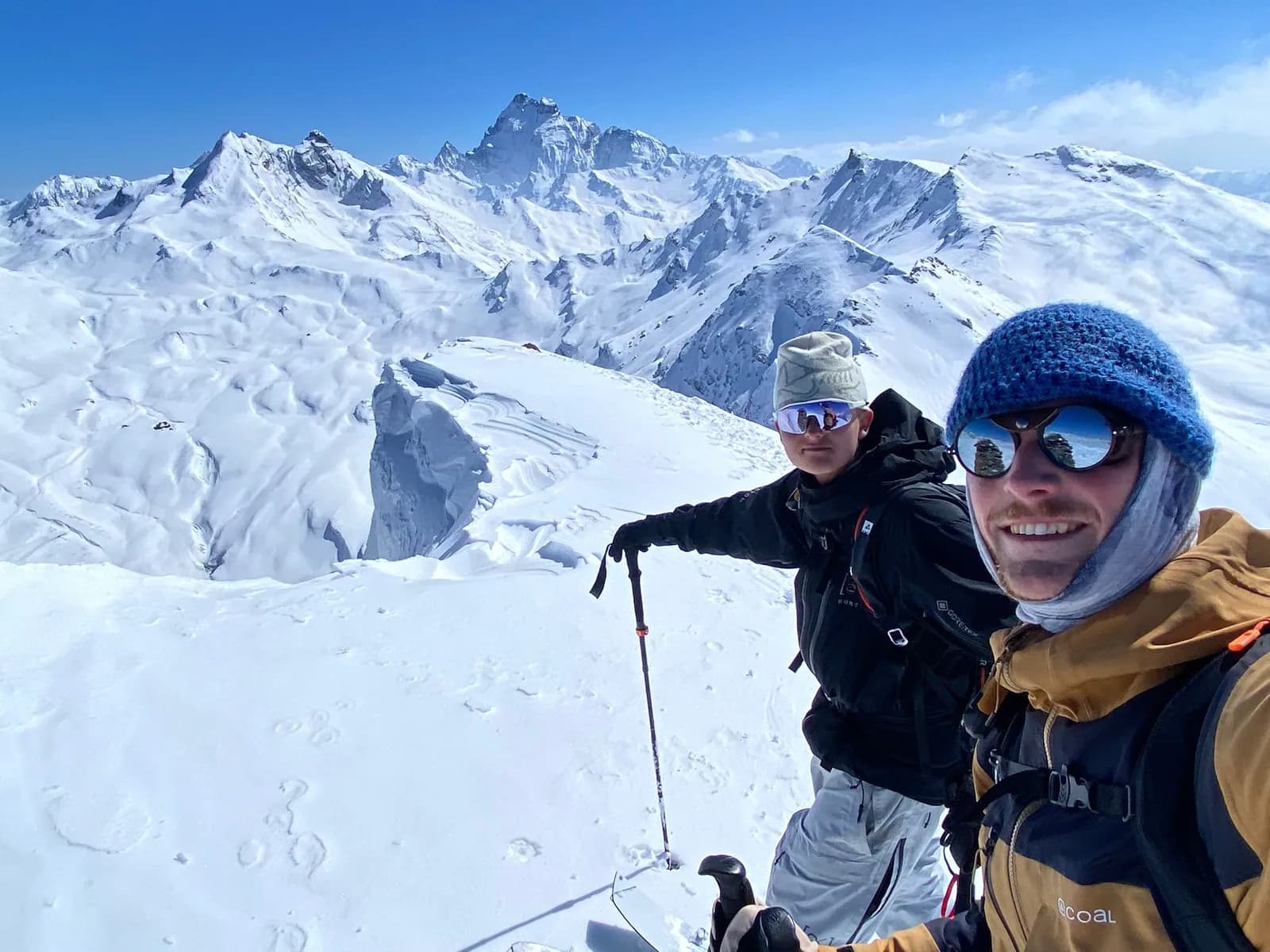 Ziggy, in front with the blue beanie, and Luuk behind him on a snowy summit ridge with poles and splitboards, a chain of peaks behind them