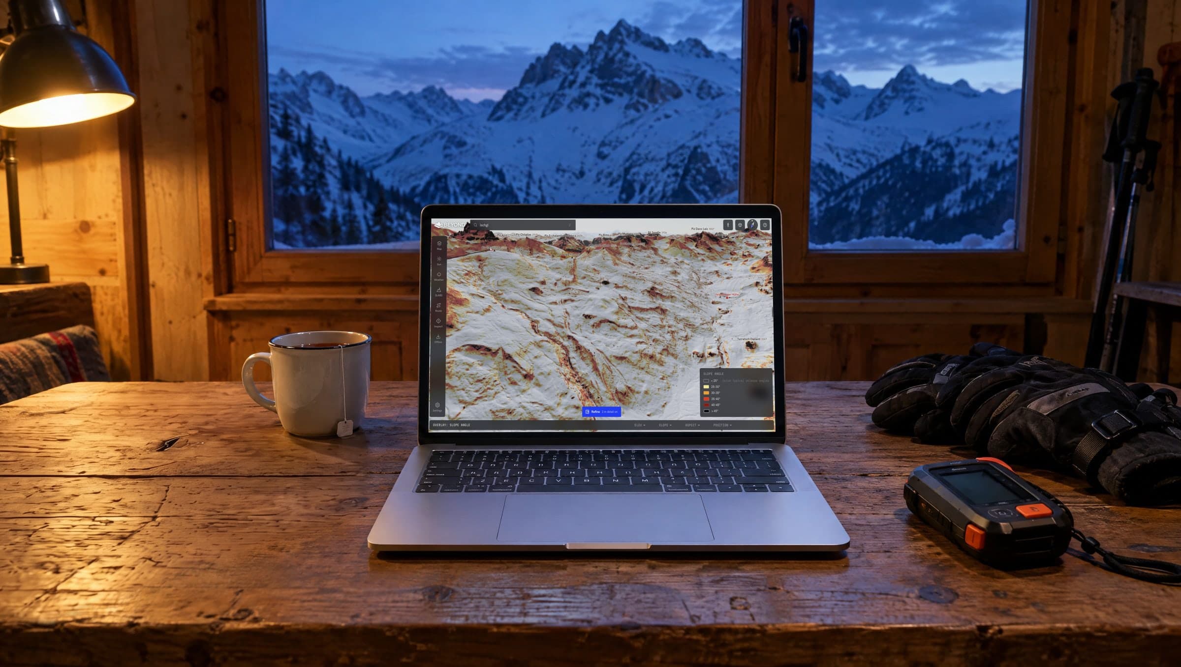 Laptop on a wooden table in a mountain hut at dusk, showing the BEYOND map with the slope-angle layer around the Heidelberger Hütte near Ischgl, next to tea, gloves and a transceiver
